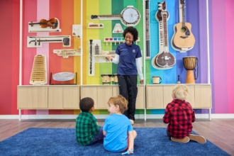 A teacher stands in front of a wall with colorful stripes and musical instruments. Children sit on the floor in front of her.